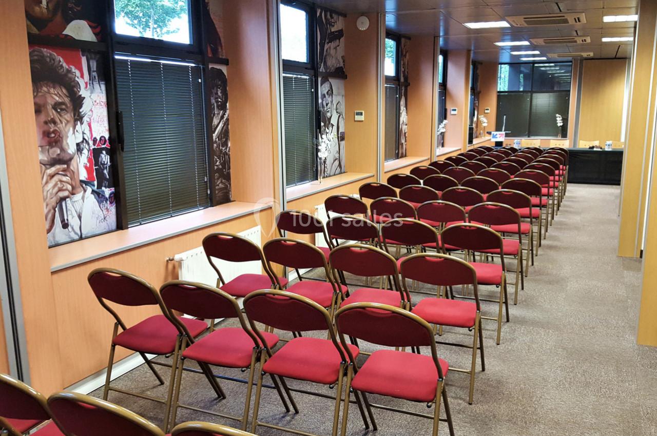 Salle de conférence avec des rangées de chaises rouges alignées devant des fenêtres et des murs décorés.