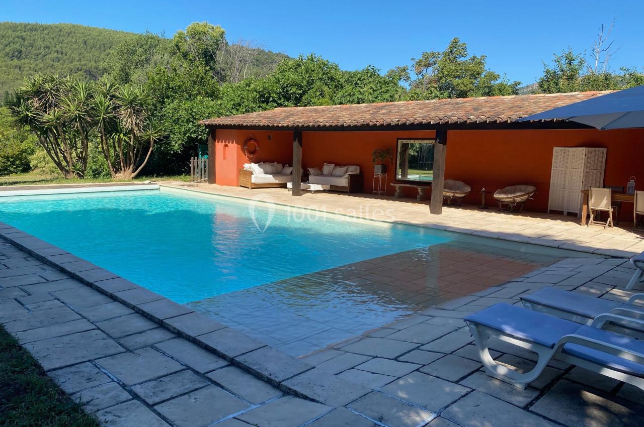 Piscine extérieure entourée de dalles en pierre, avec une maison à façade rouge et un paysage boisé en arrière-plan.
