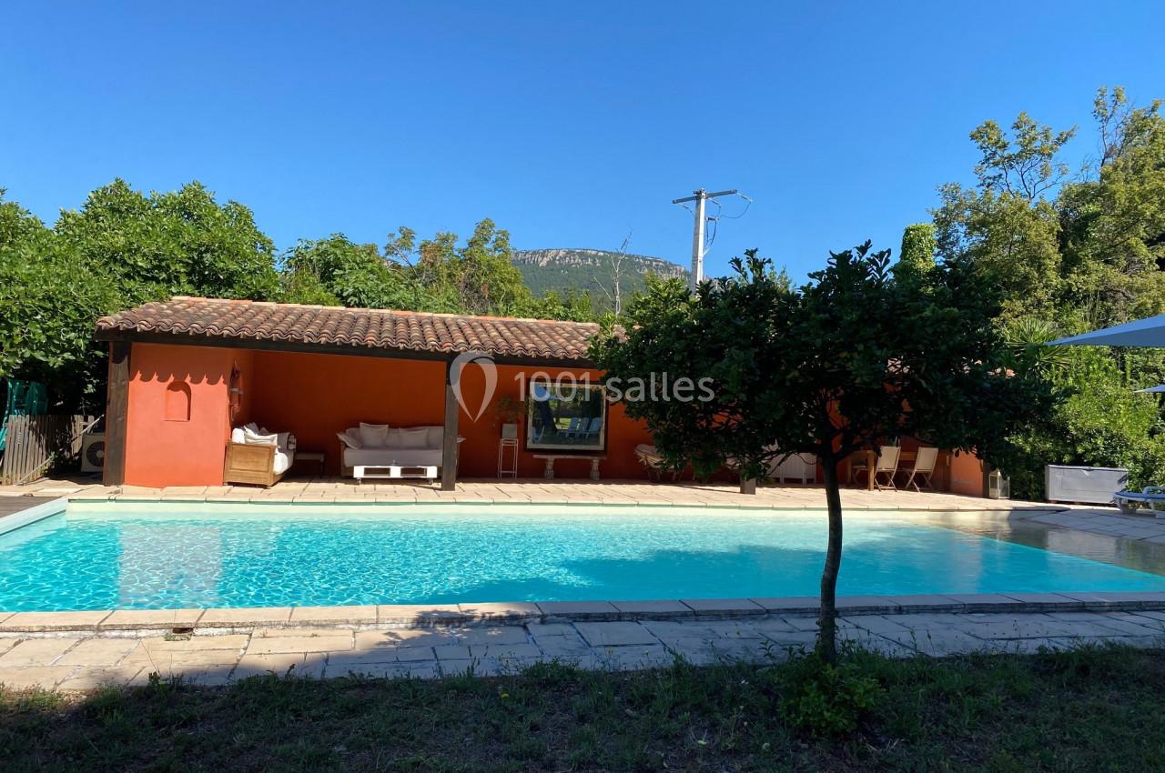 Maison avec façade orange, piscine turquoise au premier plan, terrasse ombragée et arbres environnants sous un ciel bleu.