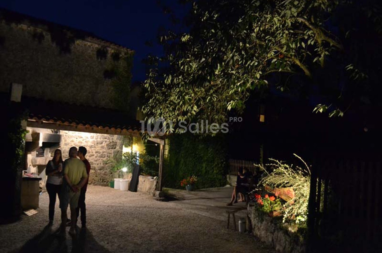 Cour extérieure éclairée de nuit avec un arbre, des plantes et trois personnes discutant près d'une maison en pierre.