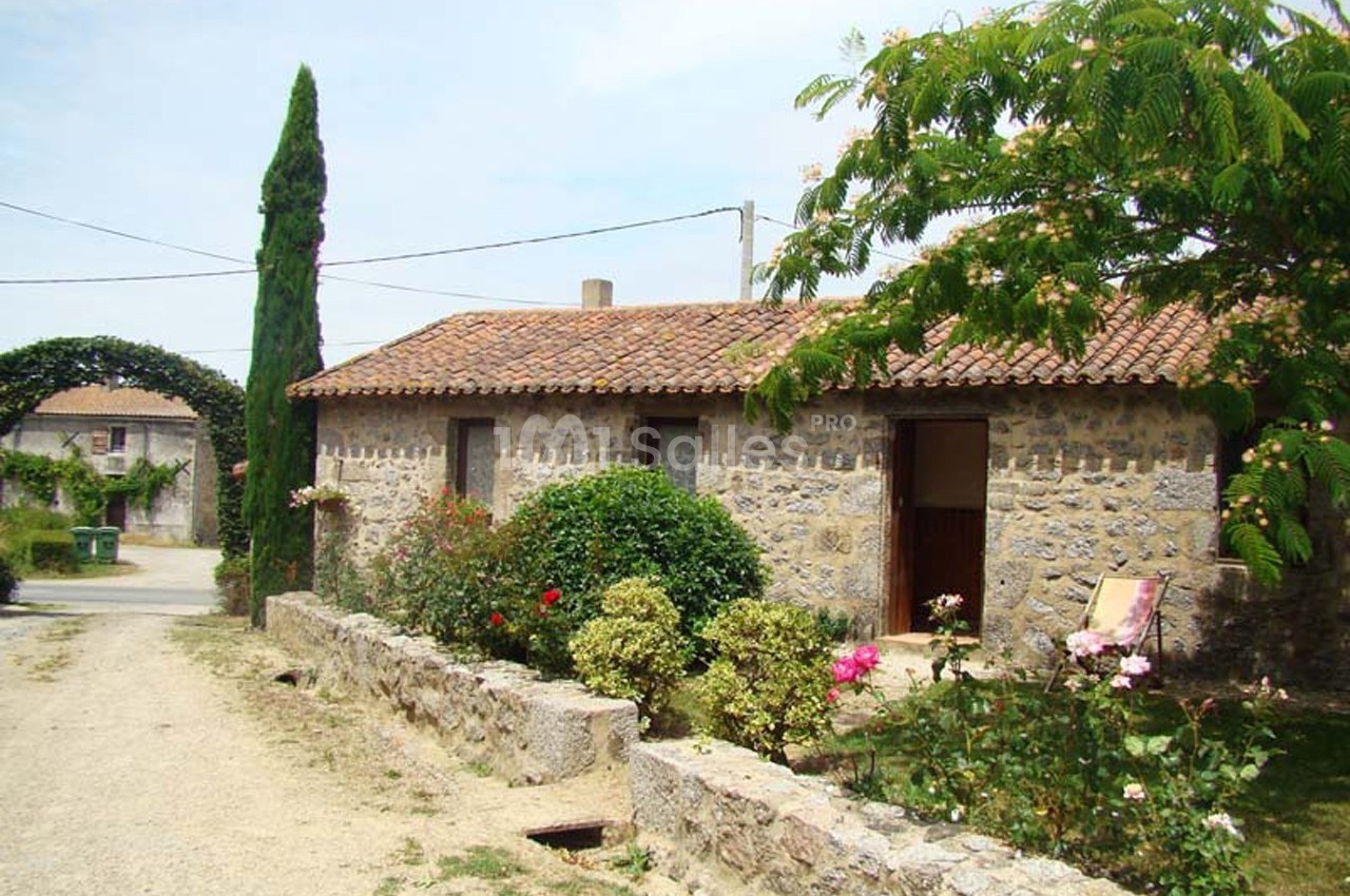 Maison en pierre avec toit en tuiles, entourée de fleurs et d'arbustes, située dans un cadre rural.