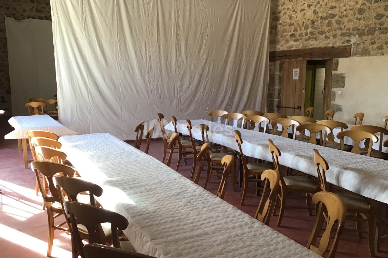 Salle avec des tables alignées et recouvertes de nappes blanches, entourées de chaises en bois, éclairée par la lumière…