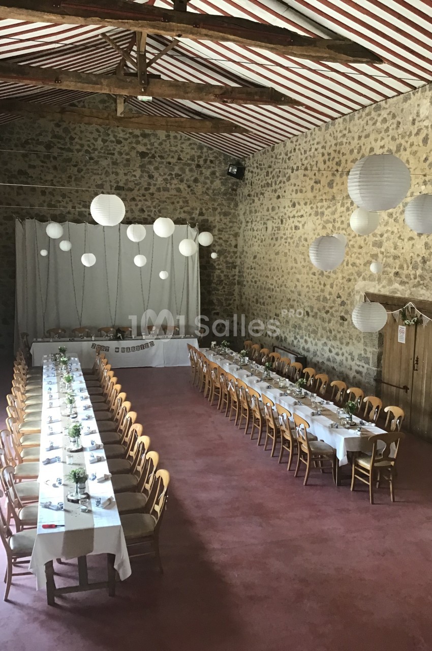 Salle en pierre avec deux longues tables dressées pour un repas, décorée de lanternes suspendues et d'éléments naturels.