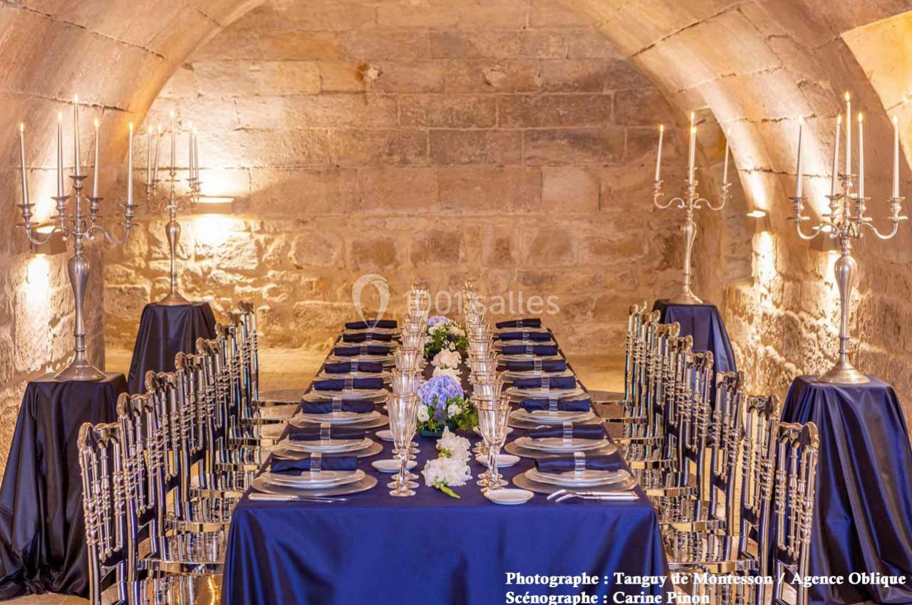 Table élégamment dressée dans une salle voûtée en pierre, ornée de chandeliers, fleurs blanches et vaisselle raffinée.
