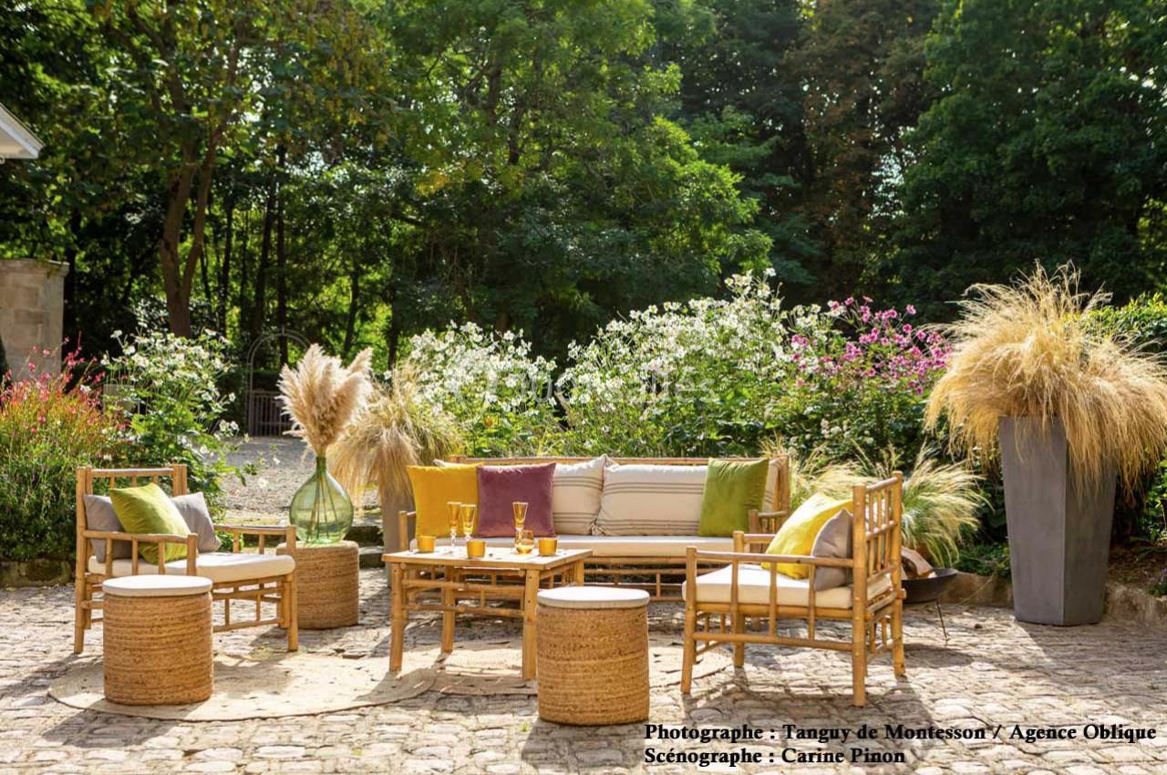 Salon de jardin en rotin avec coussins colorés, entouré de végétation et de fleurs, sur une terrasse pavée.