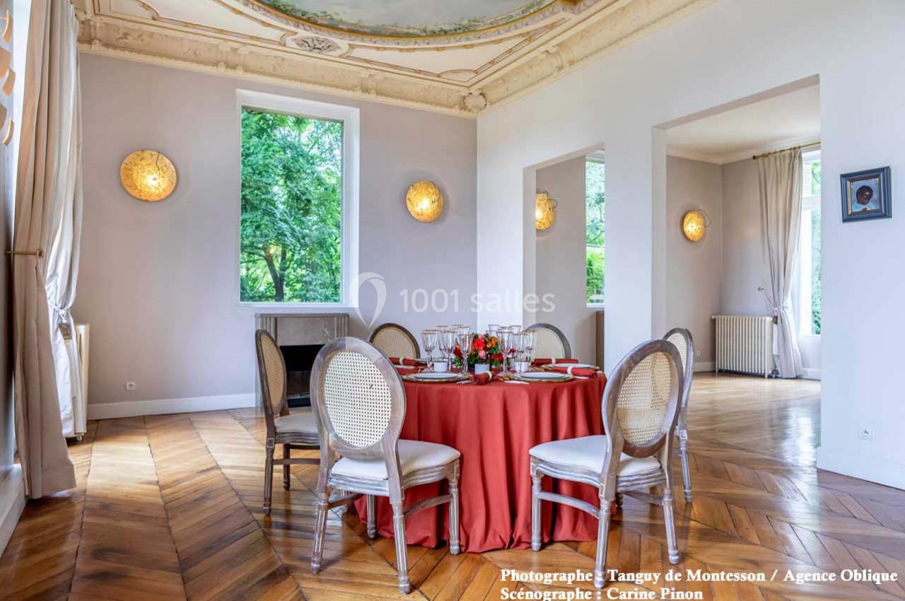 Salle lumineuse avec parquet en bois, table ronde dressée avec nappe rouge, et moulures décoratives au plafond.