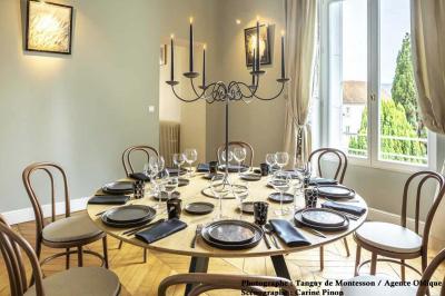 Salle lumineuse avec parquet en bois, table ronde dressée avec nappe rouge, et moulures décoratives au plafond.