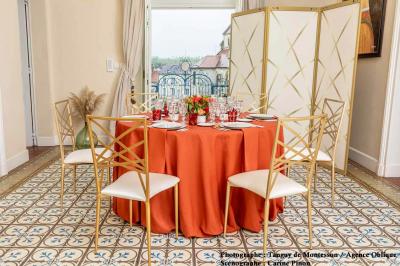 Salle lumineuse avec parquet en bois, table ronde dressée avec nappe rouge, et moulures décoratives au plafond.