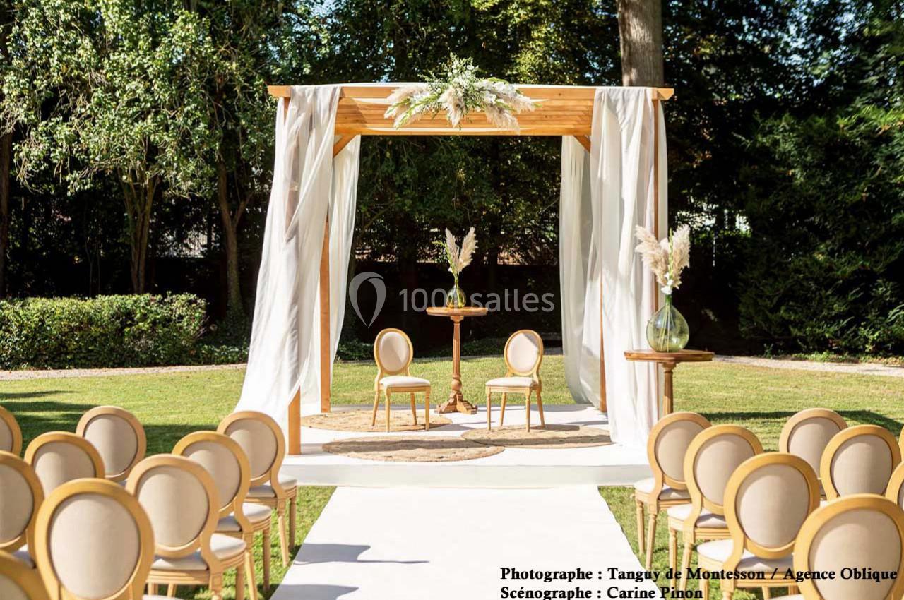 Structure en bois décorée de fleurs blanches, entourée de chaises beige, installée dans un jardin pour une cérémonie.