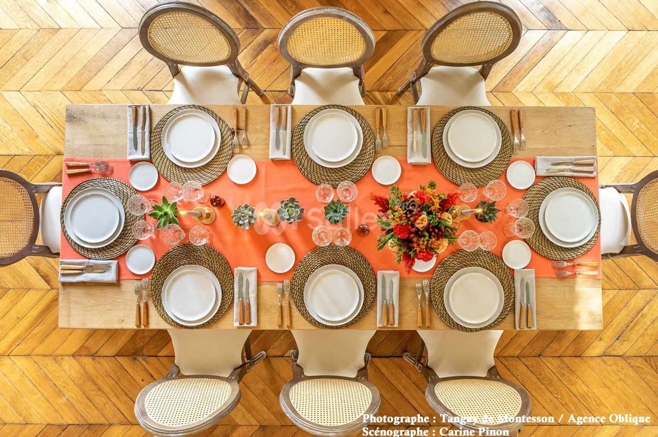 Table dressée avec nappes orange, assiettes blanches, couverts, verres et un centre de table floral coloré.