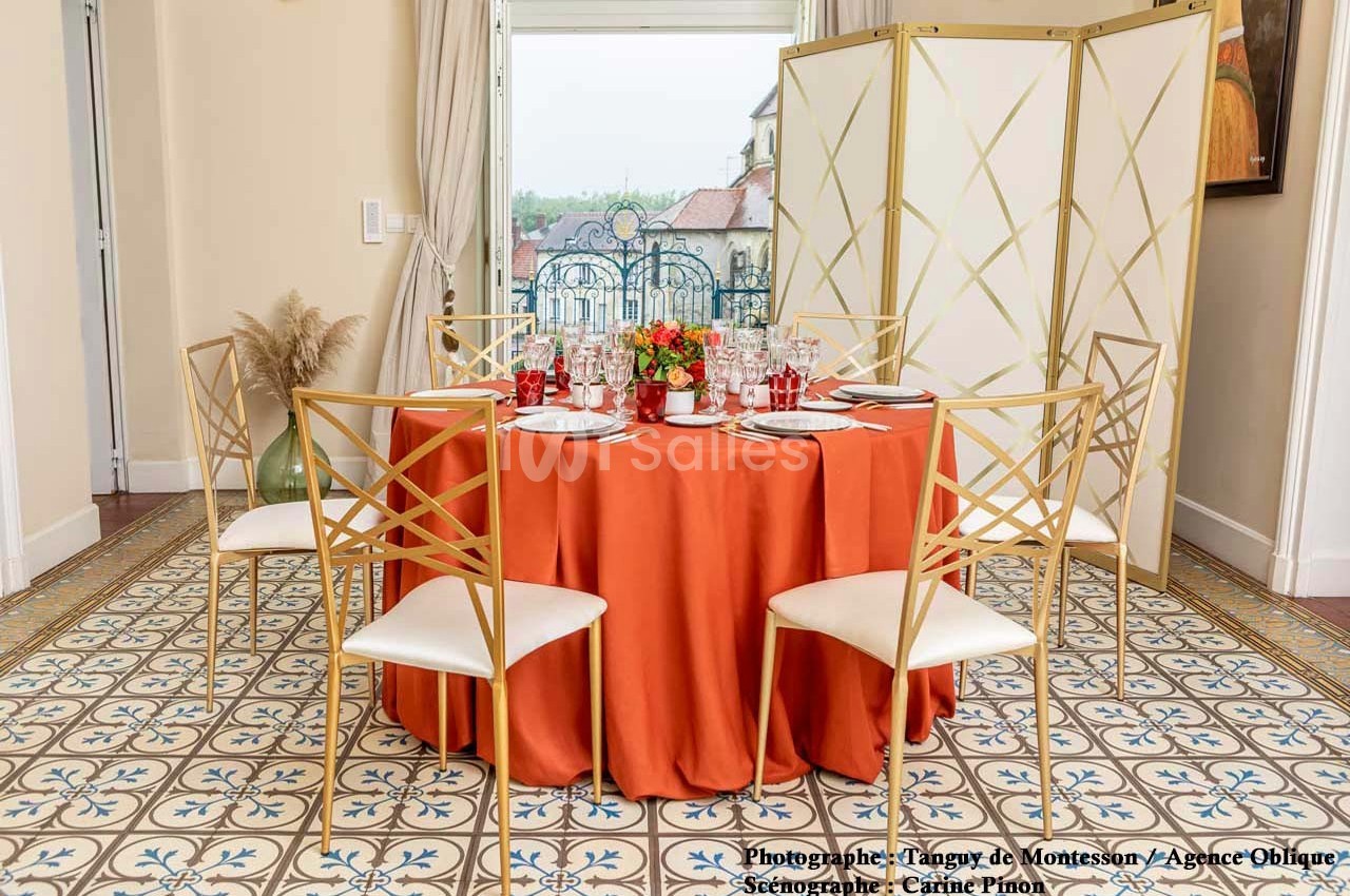 Table dressée avec une nappe orange, chaises dorées, vaisselle élégante et paravent en arrière-plan dans une pièce lumineuse.