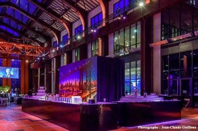 Salle lumineuse avec parquet en bois, table ronde dressée avec nappe rouge, et moulures décoratives au plafond.