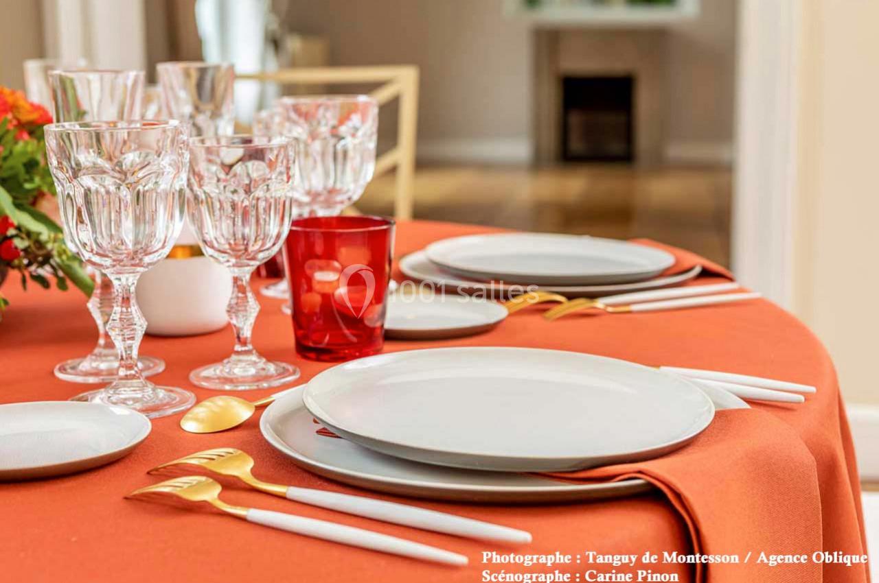 Table dressée avec une nappe orange, vaisselle blanche, couverts dorés, verres en cristal et un verre rouge.