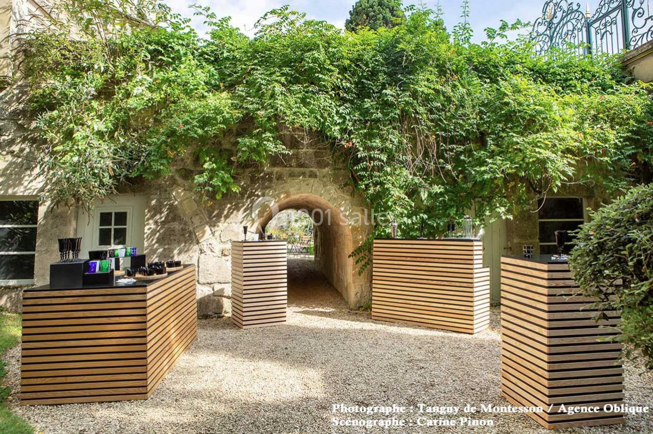 Allée extérieure avec arches en pierre, entourée de végétation, et présentoirs en bois alignés sur du gravier.