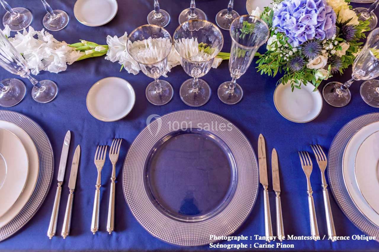 Table dressée avec vaisselle élégante, couverts en argent, verres à pied et centre de table floral sur une nappe bleue.