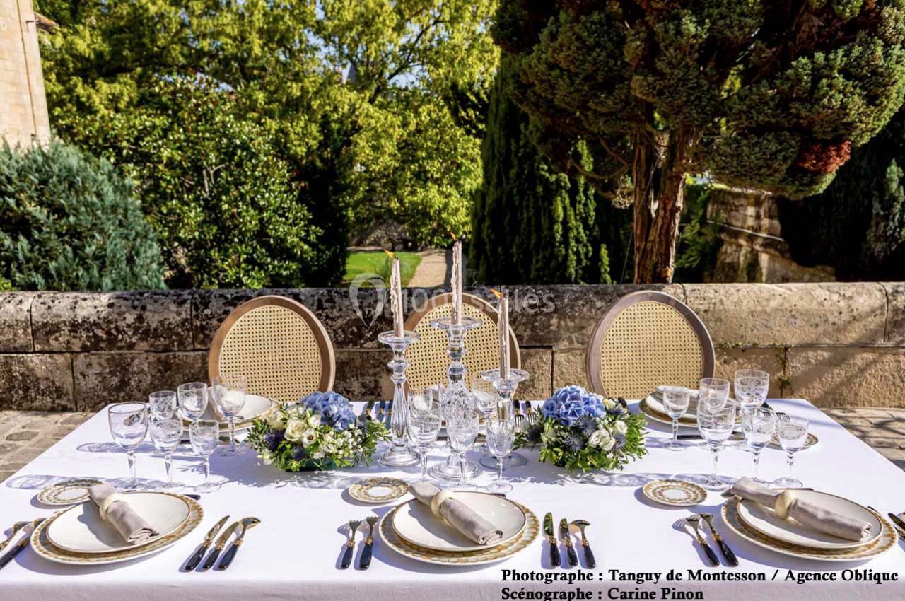 Table élégamment dressée en extérieur avec vaisselle raffinée, compositions florales et vue sur un jardin arboré.