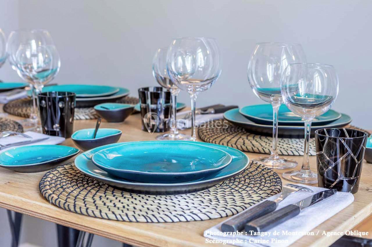 Table dressée avec des assiettes turquoise, des verres à pied, des couverts et des accessoires noirs sur un chemin de table…
