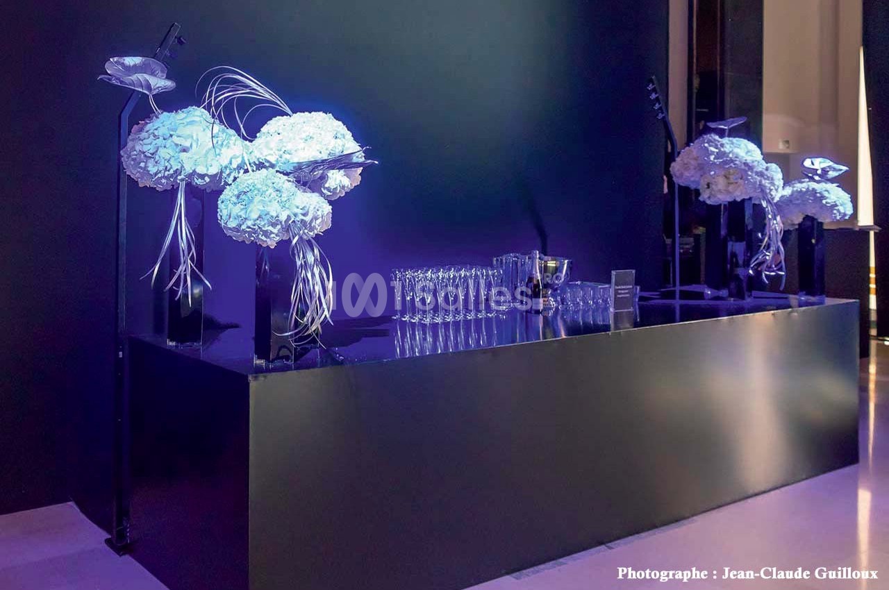 Table noire élégante décorée de fleurs blanches et de verres alignés, éclairée par une lumière violette.