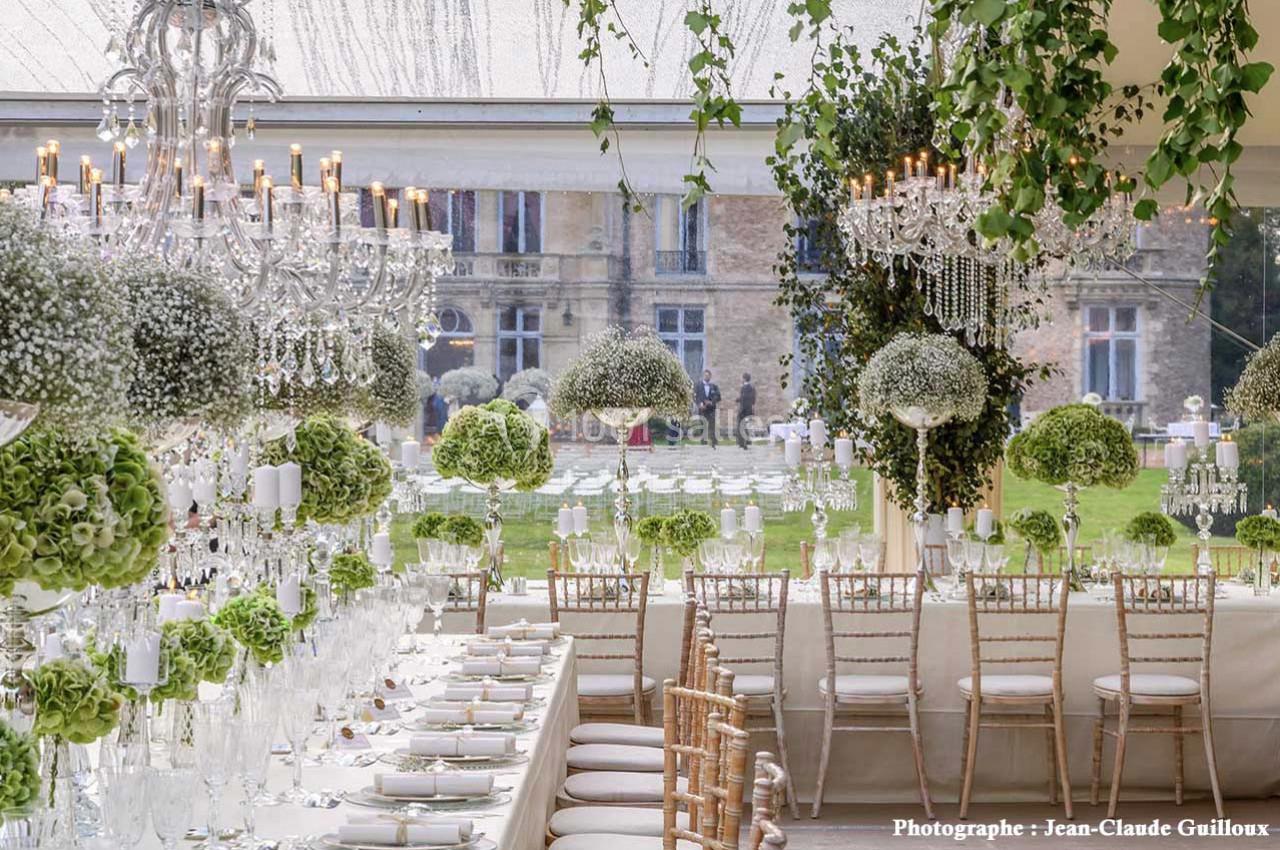Salle de réception élégante avec tables décorées de fleurs blanches et vertes, chandeliers et vue sur un jardin.