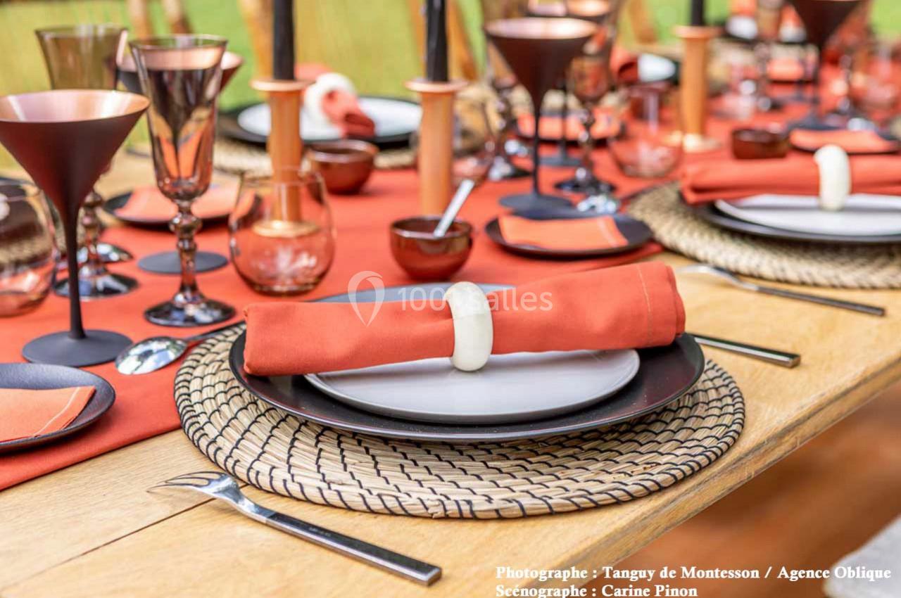 Table dressée avec des assiettes noires, serviettes orange nouées, verres cuivrés et décorations en bois sur une nappe rouge.