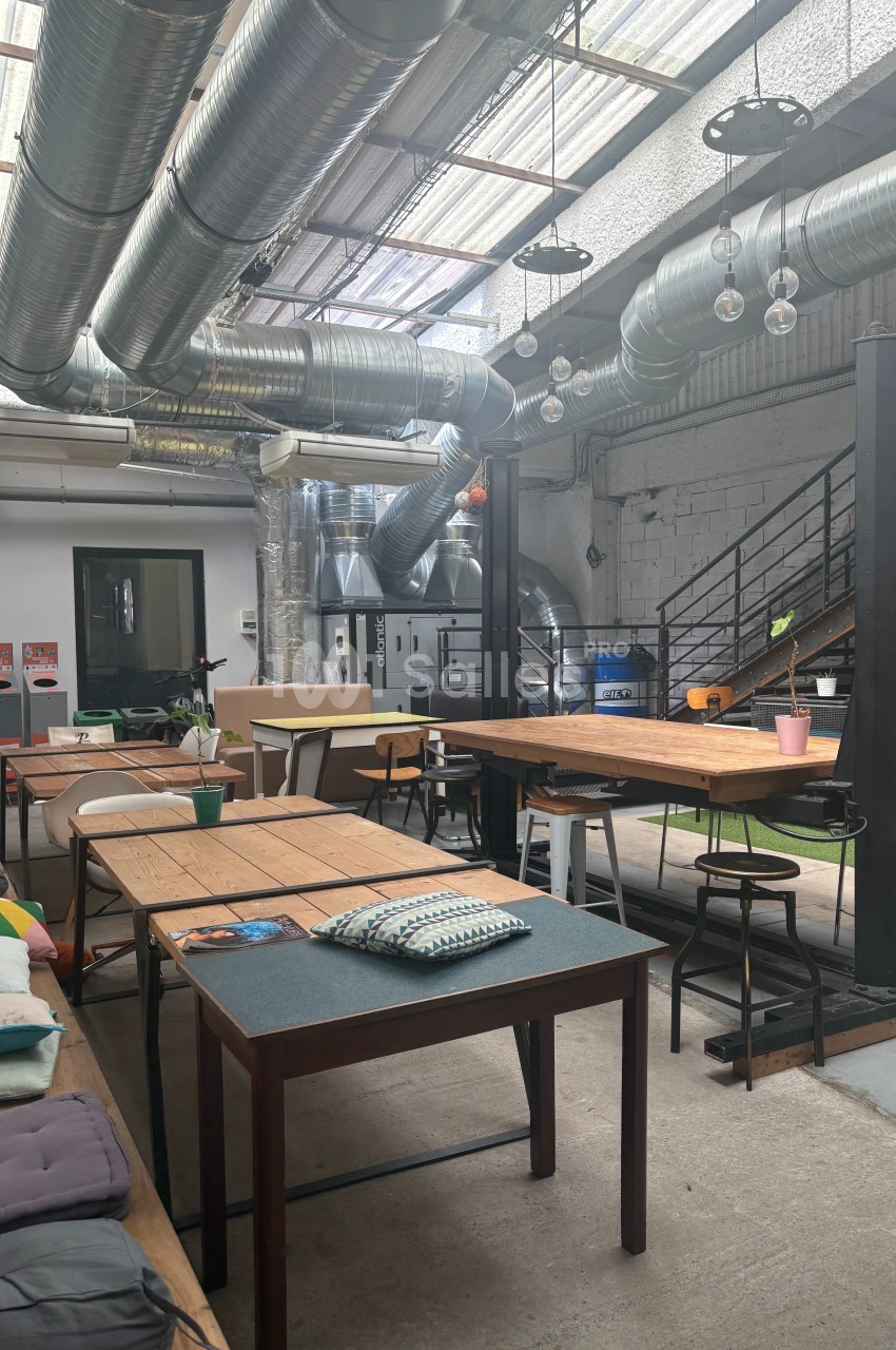 Espace de coworking lumineux avec des tables en bois, des chaises variées et un plafond industriel avec des conduits…