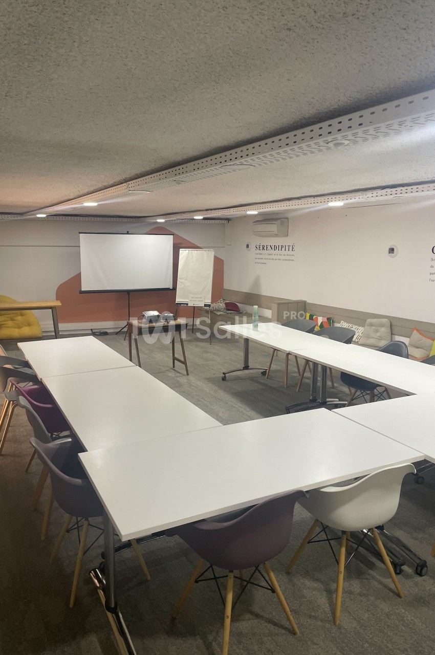 Salle de réunion avec tables disposées en U, chaises colorées, projecteur, écran et décorations murales.