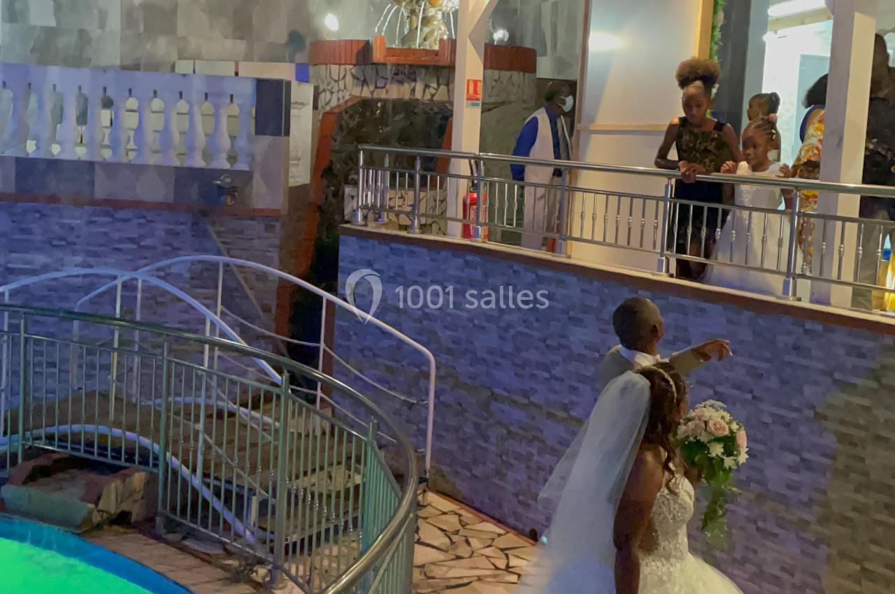 Un couple de mariés pose près d'une piscine éclairée, entouré d'invités sur une terrasse en soirée.