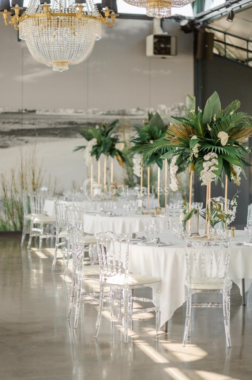 Salle de réception élégante avec tables décorées de nappes blanches, chaises transparentes et centres de table floraux.