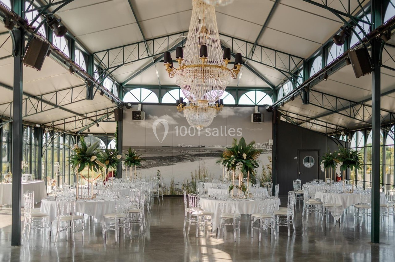 Salle de réception élégante avec tables dressées, chaises transparentes, grands lustres et décorations végétales.