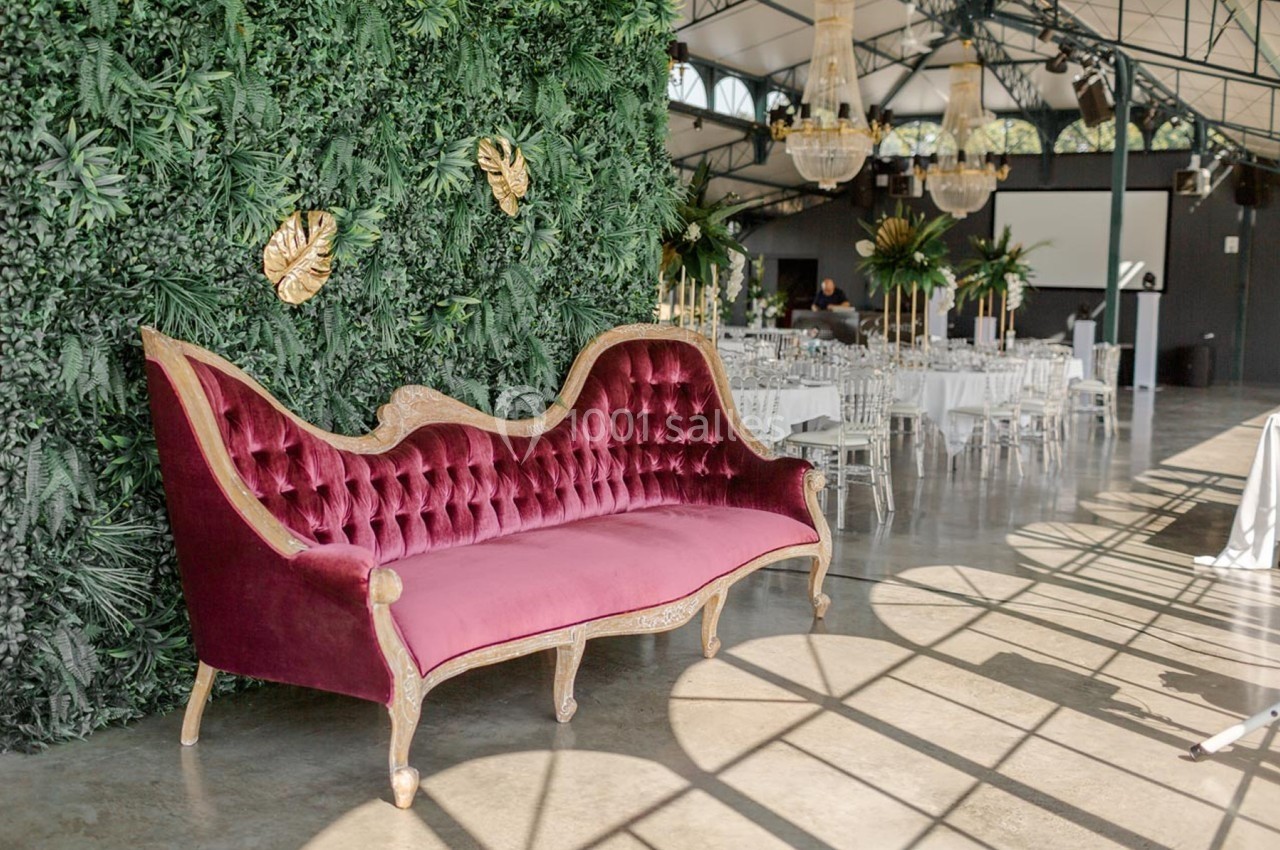 Canapé en velours rouge avec cadre doré devant un mur végétal dans une salle lumineuse décorée pour un événement.