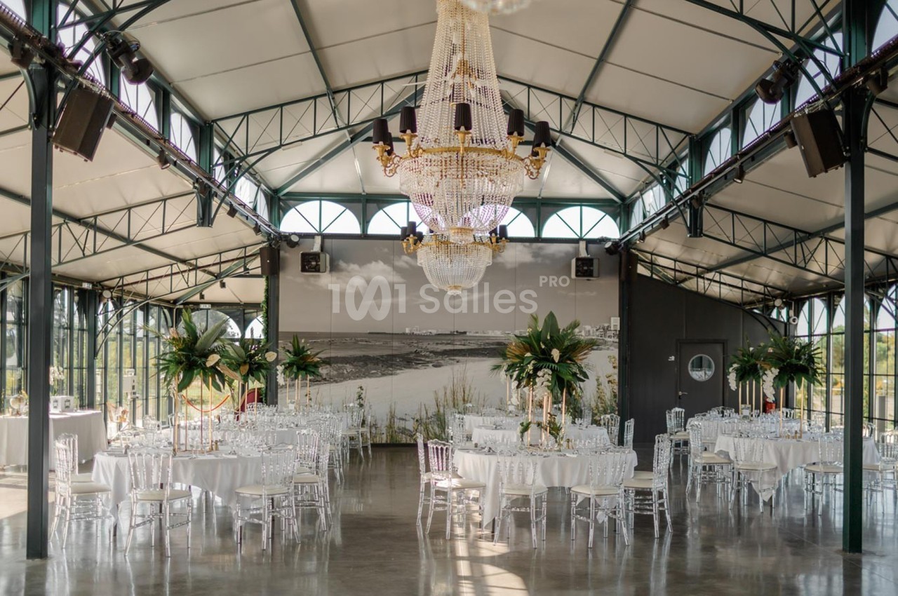 Salle de réception élégante avec tables dressées, chaises transparentes, grands lustres et décorations végétales.
