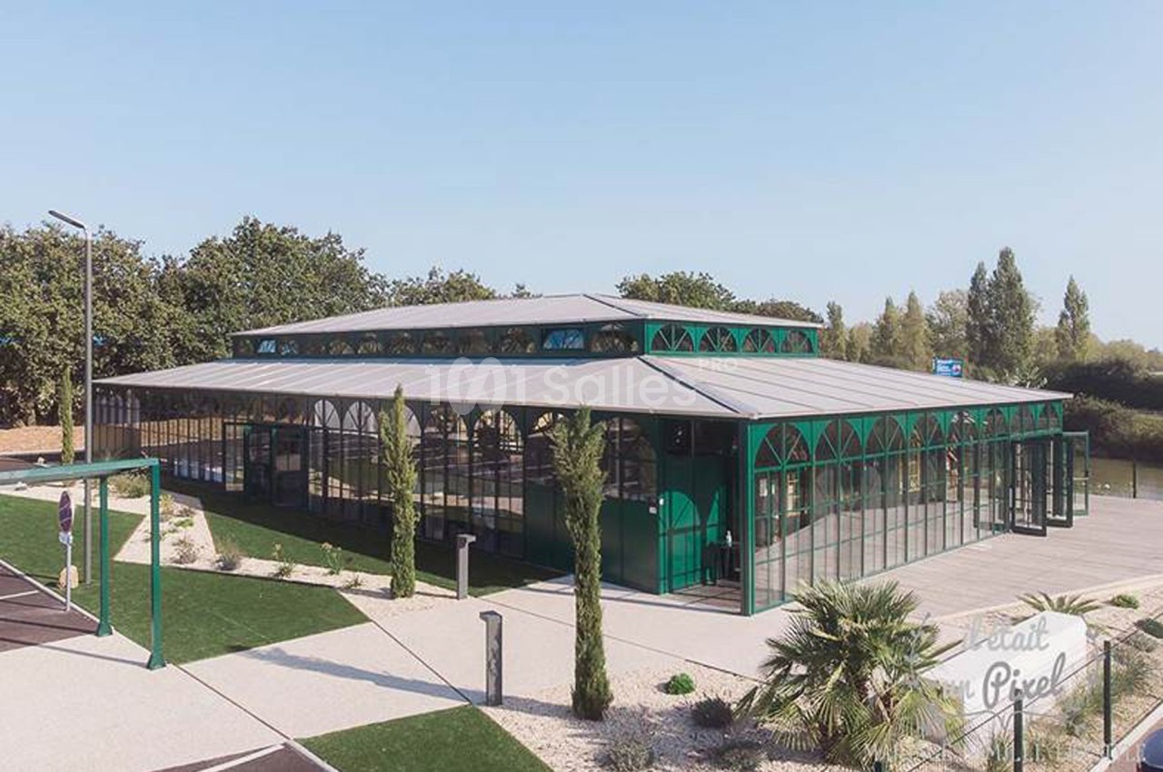 Vue extérieure d'un bâtiment moderne en verre et métal, entouré de végétation et d'un parking.
