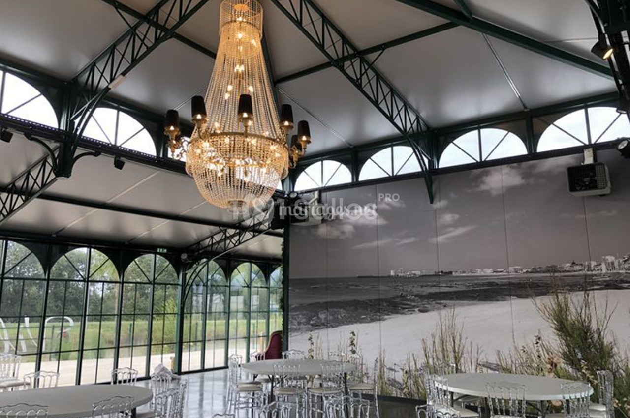 Salle lumineuse avec verrière, lustre imposant et tables rondes, décorée d'une grande photo en noir et blanc.