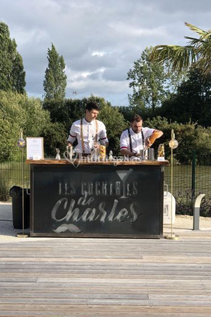 Deux barmans préparent des cocktails derrière un comptoir extérieur, entourés de verdure et d'un ciel nuageux.