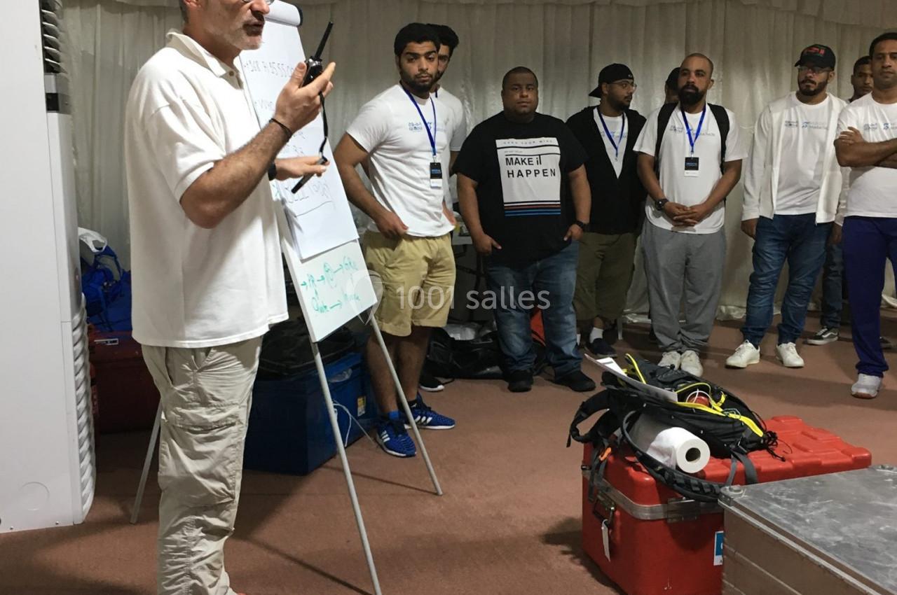 Un homme présente devant un groupe de personnes debout, dans une salle avec des équipements techniques au sol.