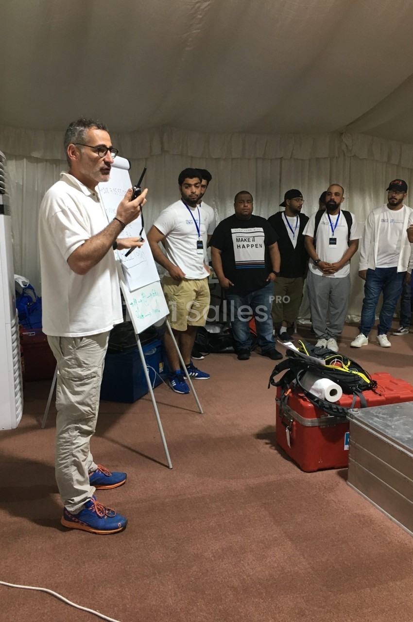 Un homme présente devant un groupe de personnes debout, dans une salle avec des équipements techniques au sol.