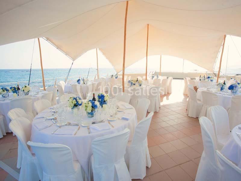 Tables rondes décorées de nappes blanches et de bouquets bleus et blancs sous une tente en bord de mer.