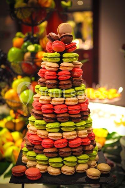 Pyramide de macarons colorés empilés sur un présentoir, entourée de fruits en arrière-plan flou.