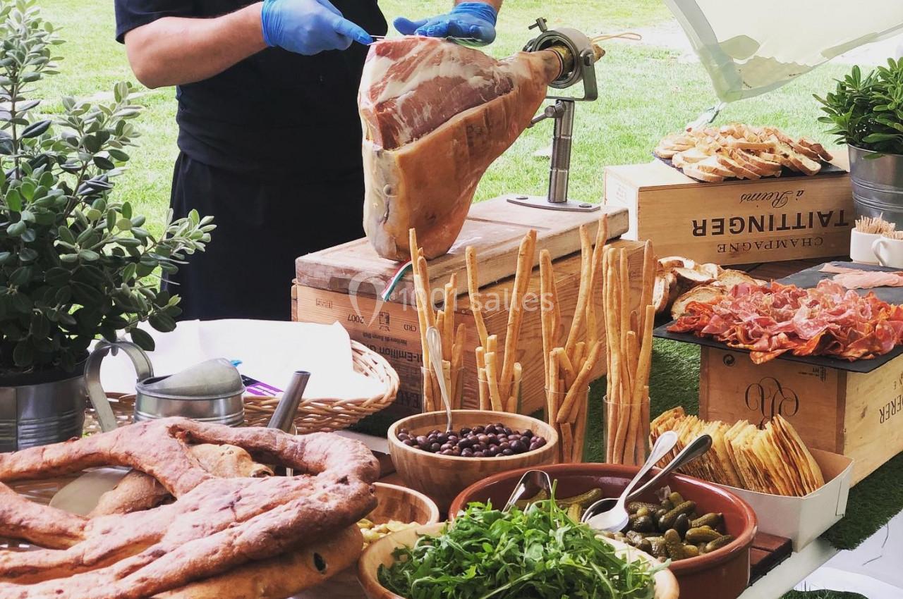 Un chef découpe un jambon cru sur un buffet garni de pains, légumes, olives et charcuteries en extérieur.