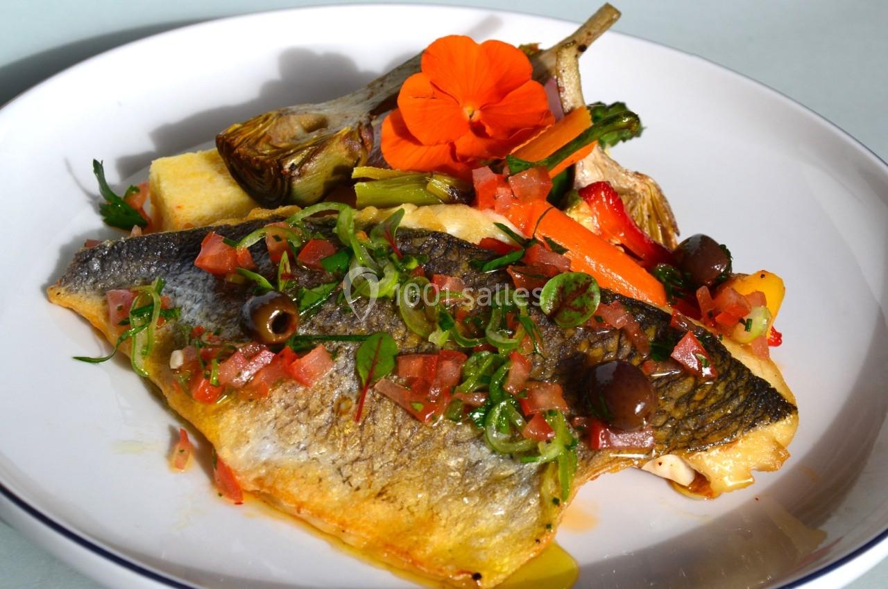 Filet de poisson grillé garni de légumes colorés, olives, herbes fraîches et une fleur comestible sur une assiette blanche.