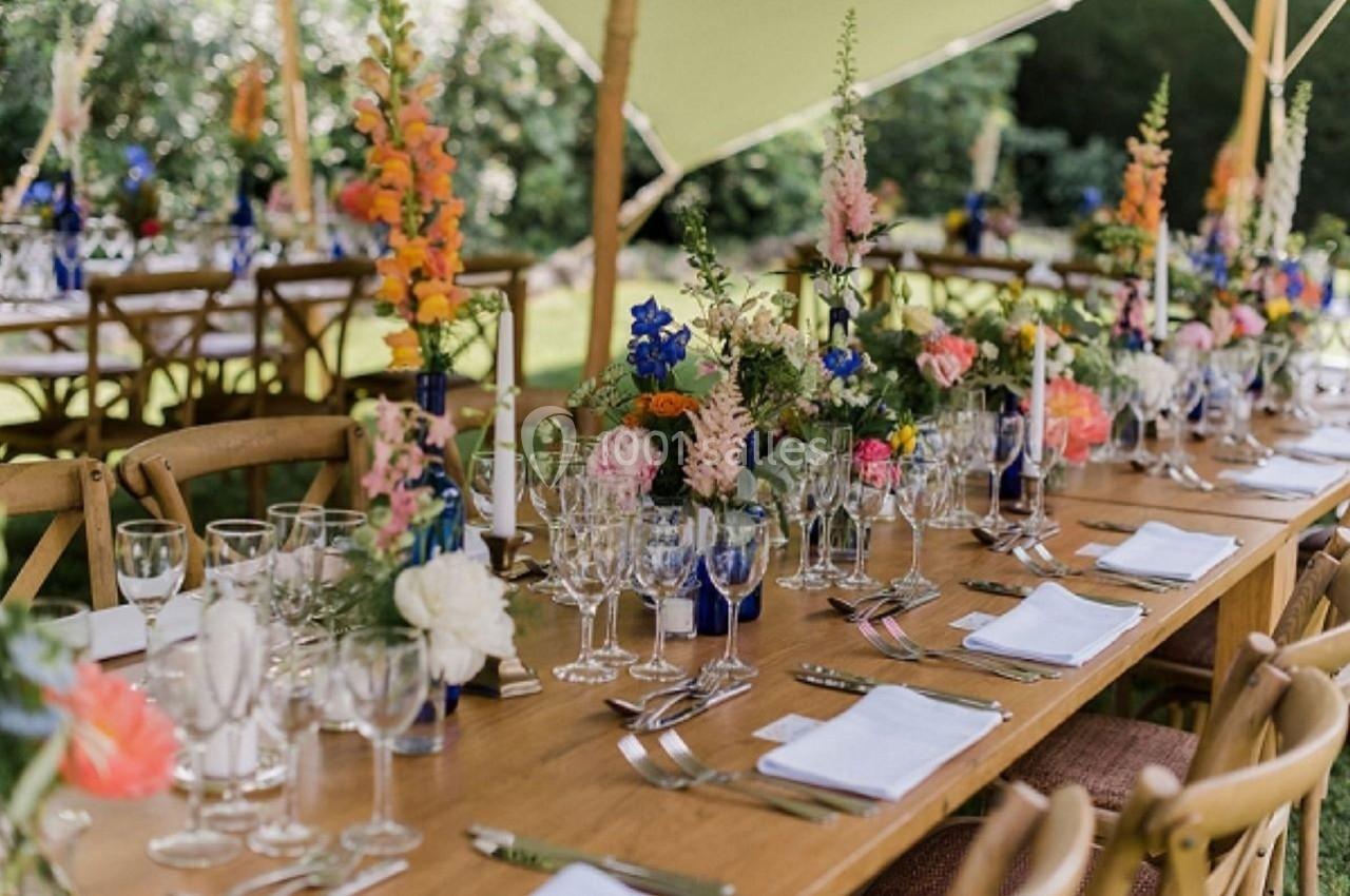Table en bois décorée de fleurs colorées, verres, assiettes et couverts, installée en extérieur sous une tente.
