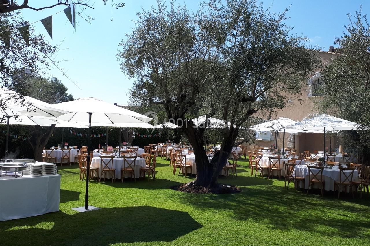 Tables dressées sous des parasols blancs dans un jardin verdoyant, entouré d'arbres et baigné de lumière naturelle.