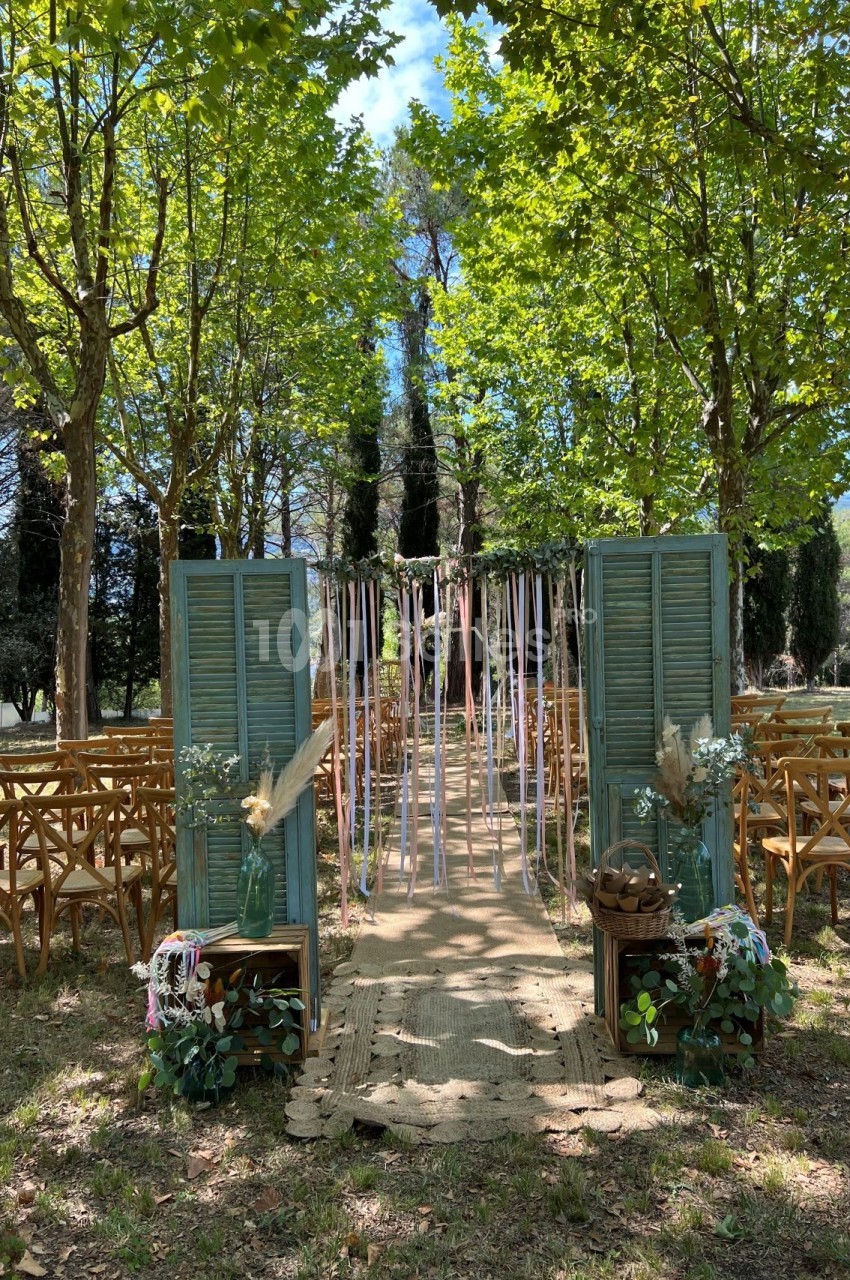 Allée décorée pour une cérémonie en plein air, entourée d'arbres, avec chaises en bois et rubans suspendus.
