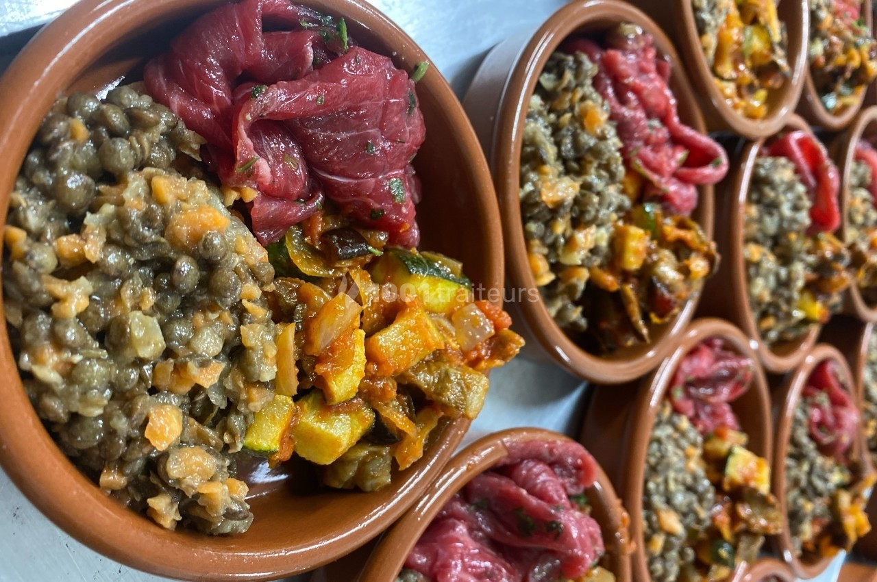 Assiettes en terre cuite contenant des lentilles, légumes sautés et morceaux de viande crue, disposées sur une table.