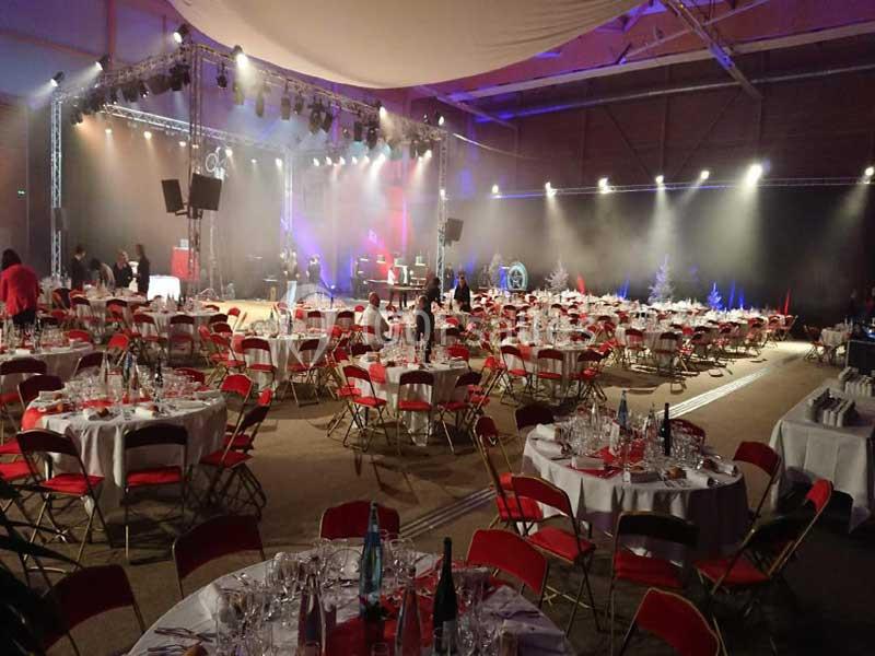 Salle de réception avec tables rondes dressées, chaises rouges et éclairage de scène en arrière-plan.