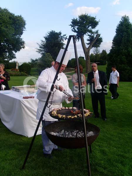 Un chef cuisine des brochettes sur un barbecue suspendu dans un jardin lors d'un événement en plein air.