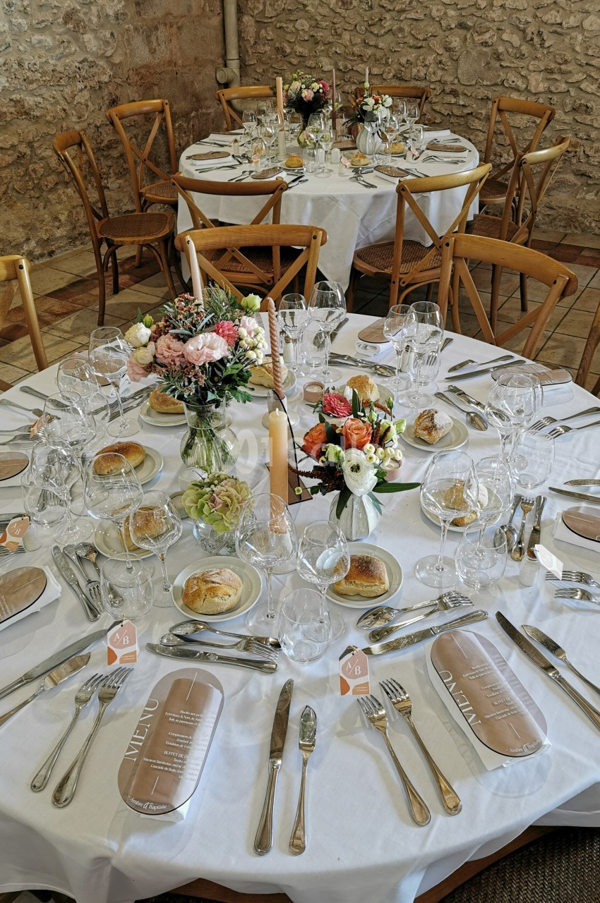 Tables rondes élégamment dressées avec nappes blanches, fleurs colorées, menus et couverts dans une salle en pierre.