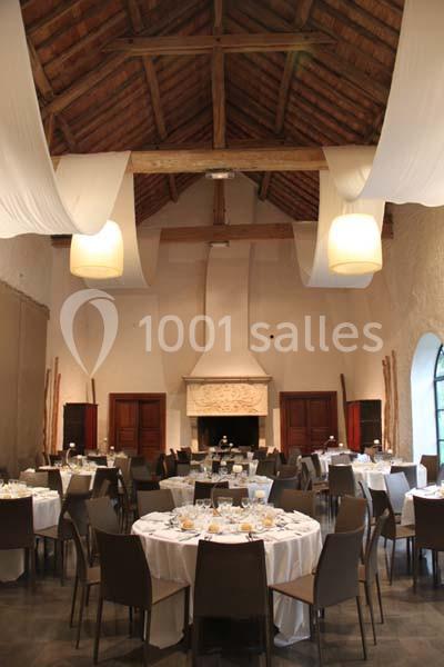 Salle de réception avec tables rondes dressées, chaises modernes, cheminée sculptée et poutres apparentes au plafond.