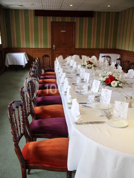 Table dressée avec nappes blanches, couverts, menus et bouquets de fleurs, entourée de chaises en bois dans une salle.