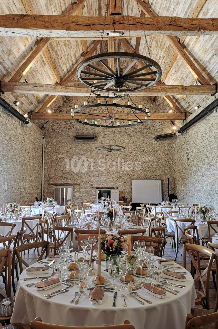 Salle de réception en pierre avec tables rondes dressées, décorations florales et poutres apparentes au plafond.